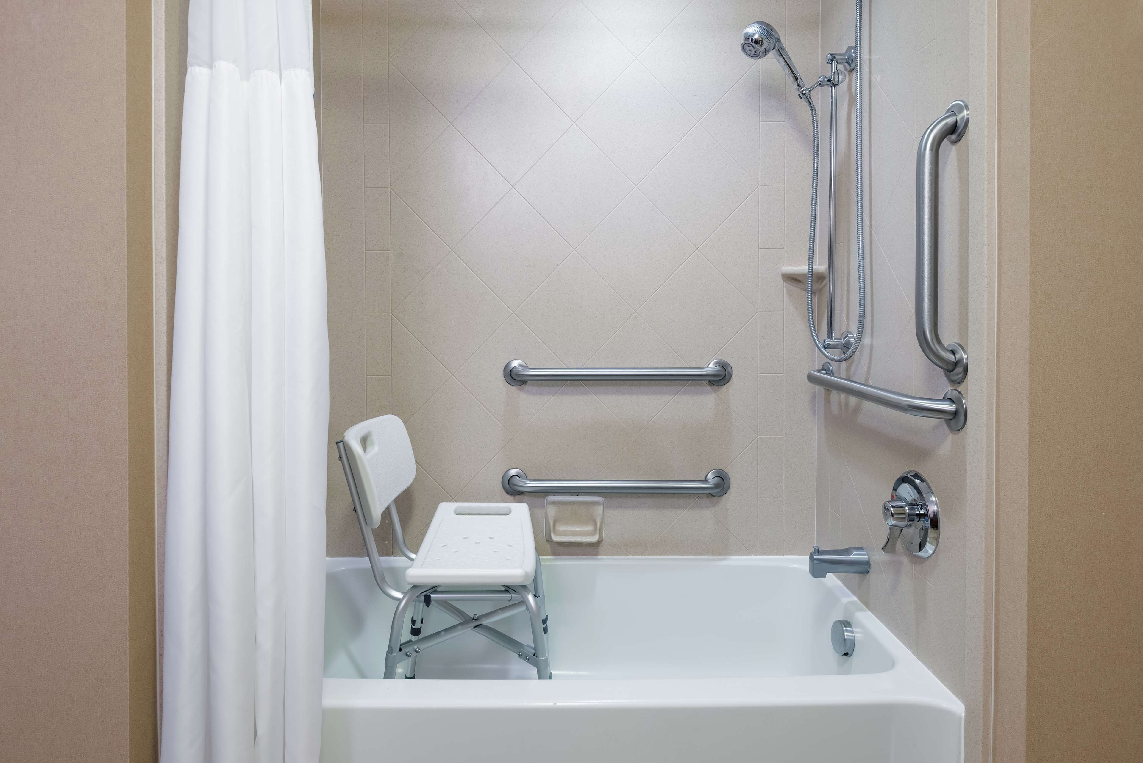 A bathroom with a white bathtub, grab bars, a shower chair, and a handheld showerhead.