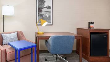 A hotel room with a blue table, a blue office chair, a desk, a lamp, a refrigerator, and a framed picture on the wall.