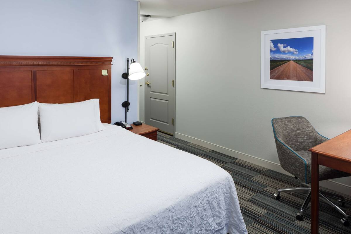 A hotel room with a large bed, a desk, a chair, a lamp, and a door.