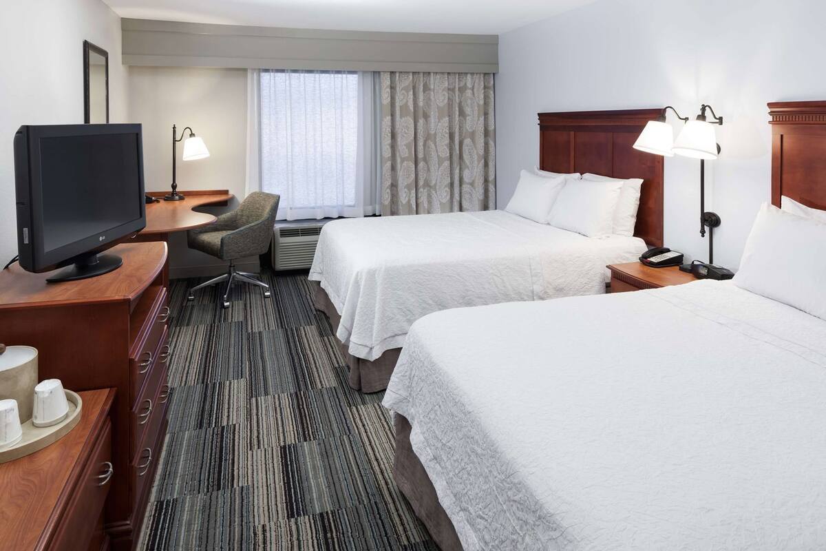 A hotel room with two beds, a desk with a TV, a chair, a desk lamp, and a bedside table with a phone.