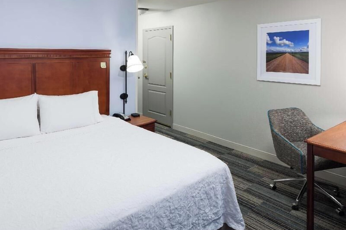 A hotel room with a bed, a desk, a chair, a lamp, and a door.