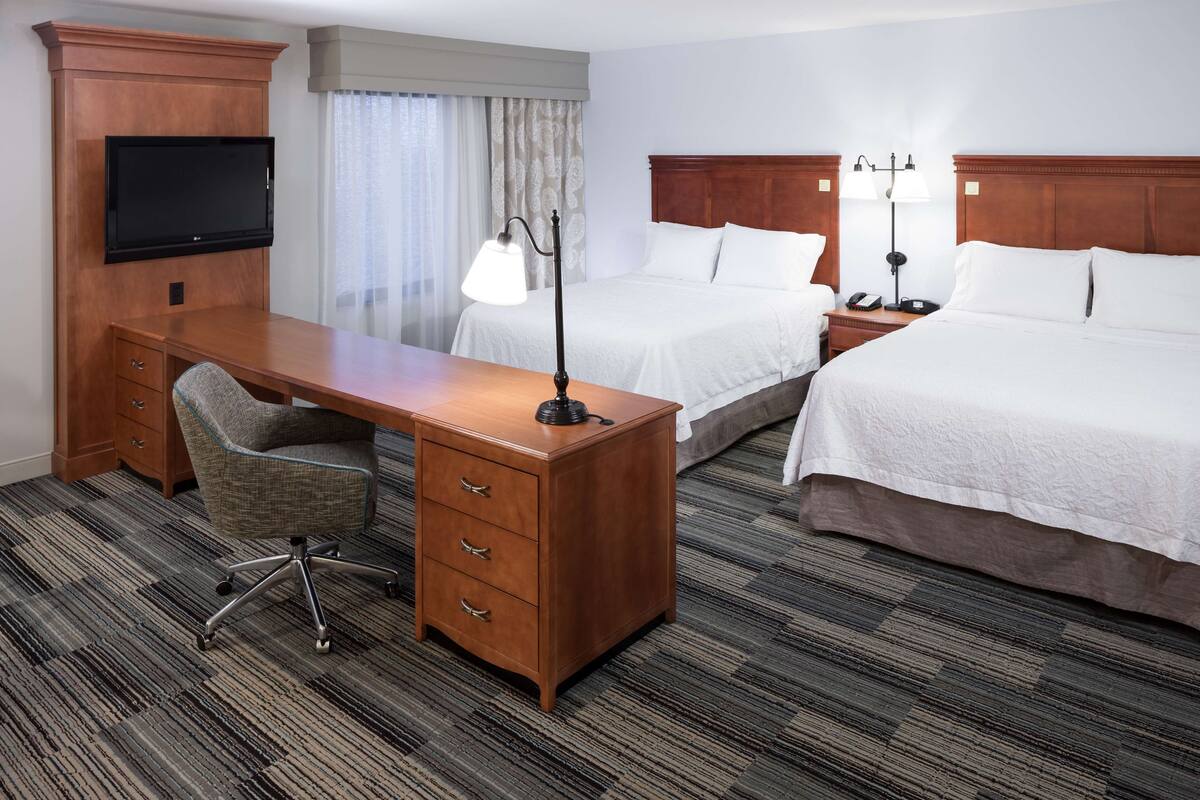 A hotel room with a wooden desk, two beds, a television, and a lamp.