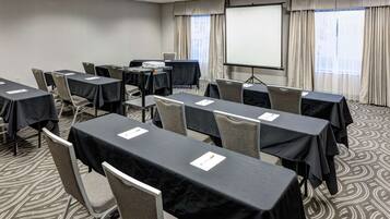 Un salón de conferencias con mesas dispuestas para una reunión, una pantalla de proyector y un amplio ventanal con cortinas.