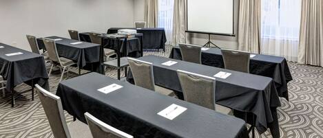 A conference room with tables set up for a meeting, a projector screen, and a large window with curtains.