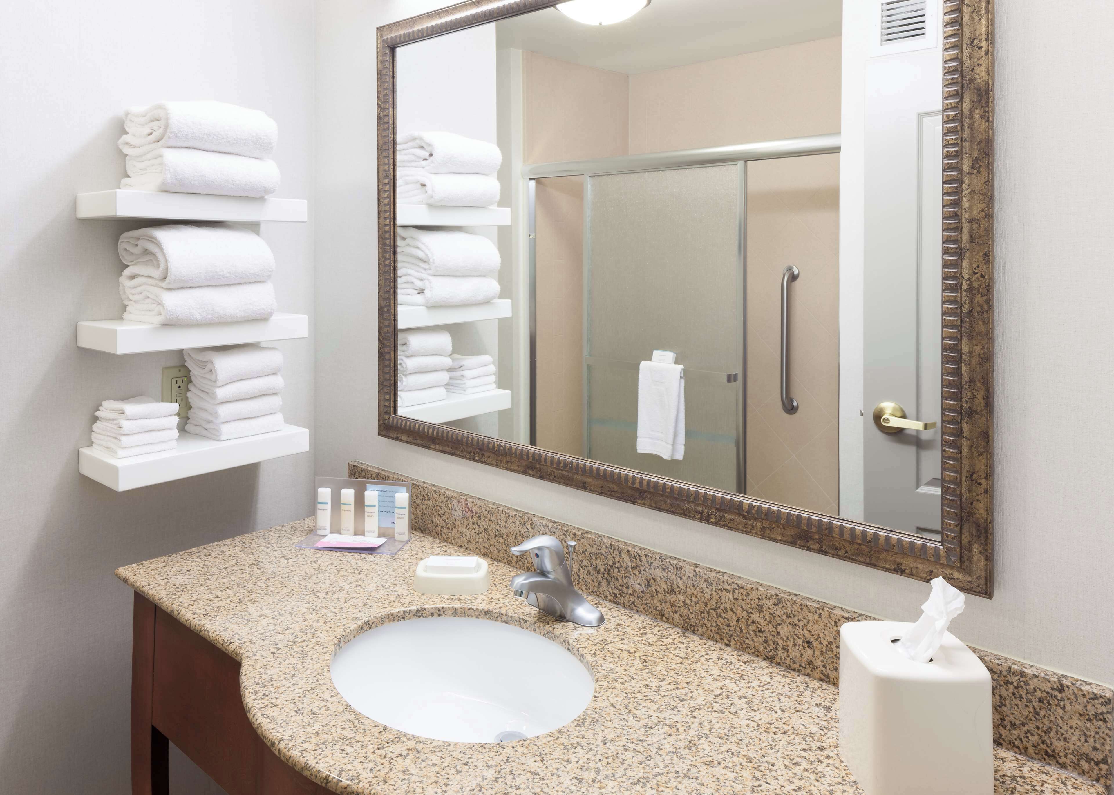 A bathroom with a granite countertop, white sink, and a large mirror. There are white towels stacked on shelves and a glass shower door.