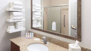 A bathroom with a granite countertop, white sink, and a large mirror. There are white towels stacked on shelves and a glass shower door.