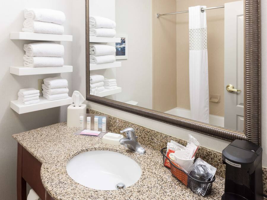 Un baño de hotel con lavamanos de encimera de granito, un espejo grande y toallas blancas dobladas ordenadamente en repisas.