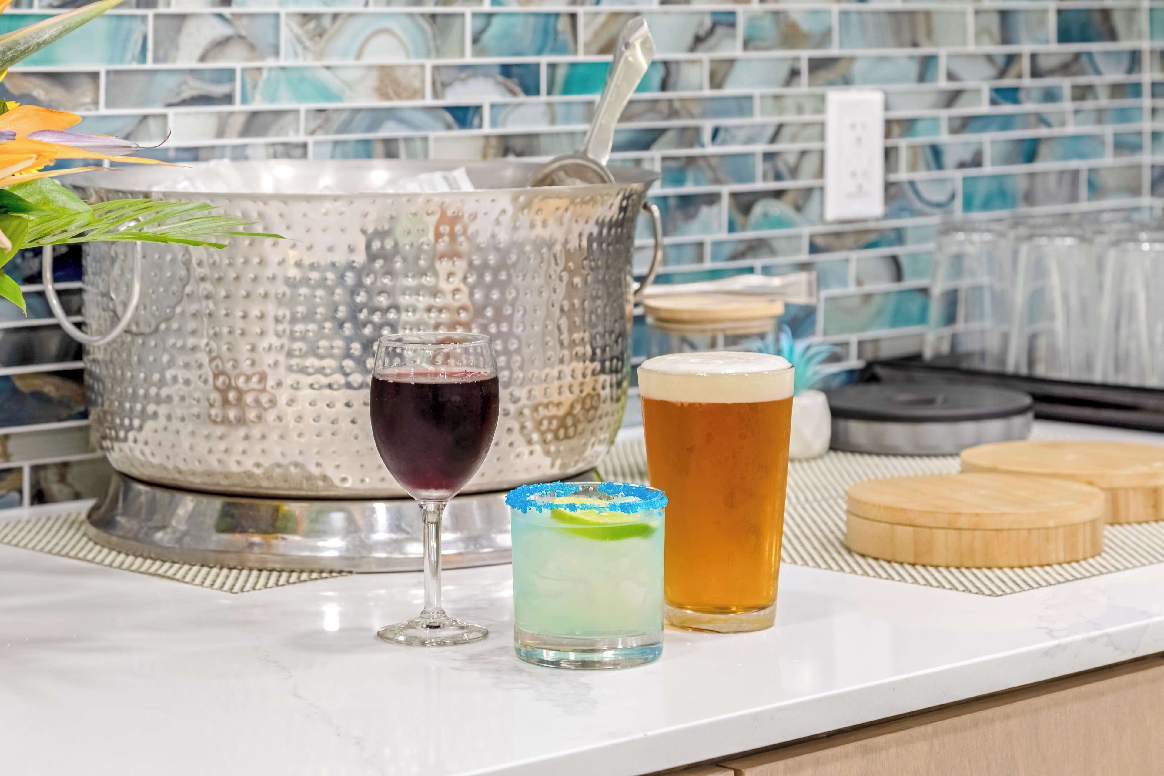 A bar area with a metal ice bucket, a glass of red wine, a cocktail with a lime garnish, and a beer on a marble countertop.