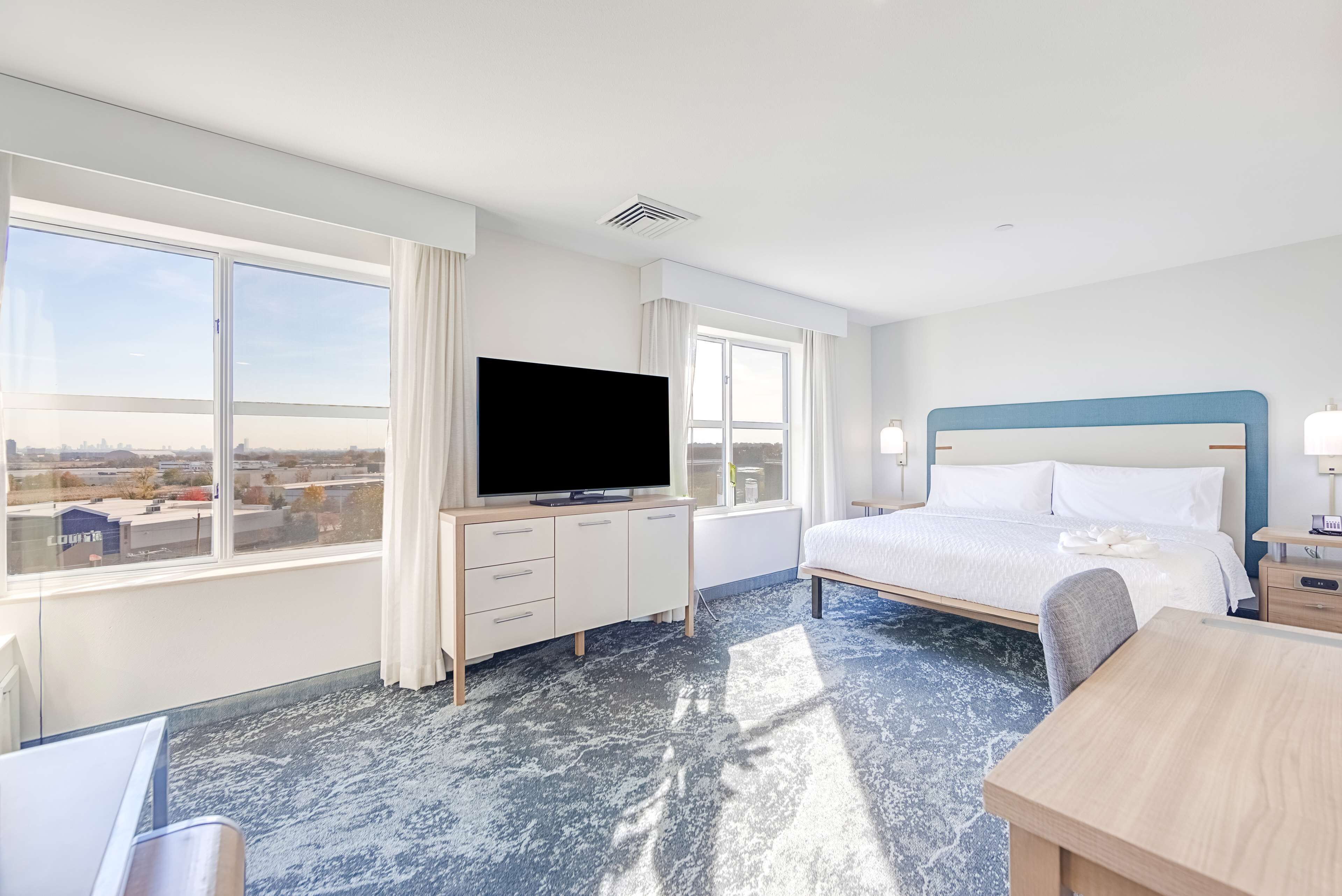Une chambre d’hôtel avec un grand lit, un bureau, une télévision et une vue sur la ville grâce à de grandes fenêtres.