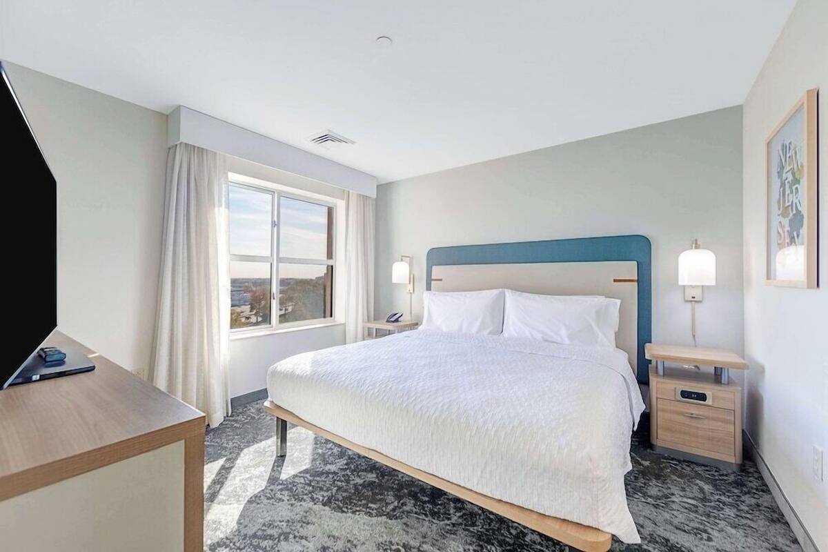 A hotel room with a bed, bedside tables, a desk, a window with curtains, and a view of the city.