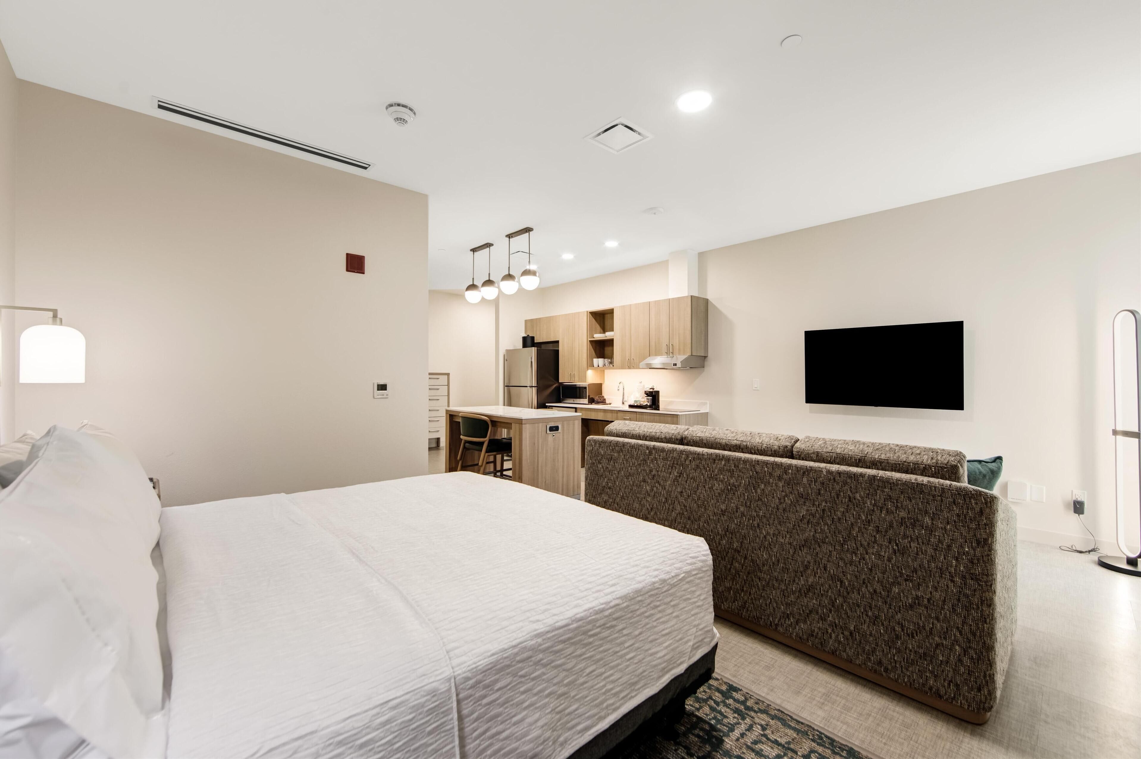 A modern hotel room with a bed, a sofa, a kitchenette, and a TV mounted on the wall.