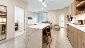 Une chambre d’hôtel moderne avec une table à manger surmontée d’un plateau en marbre, des chaises en bois, un canapé, un téléviseur à écran plat, un four encastré et un coin cuisine doté de meubles en bois et d’un plan de travail.
