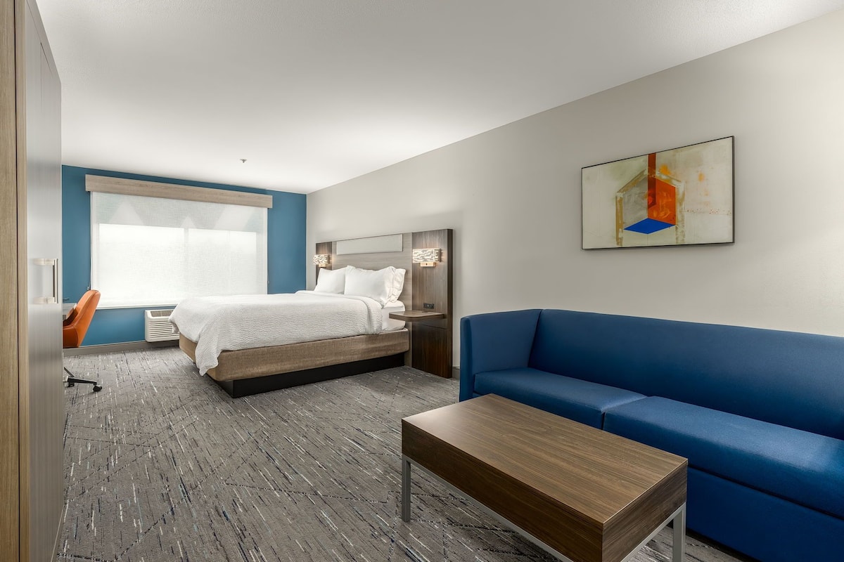 A modern hotel room with a bed, a blue sofa, a wooden coffee table, and a painting on the wall.