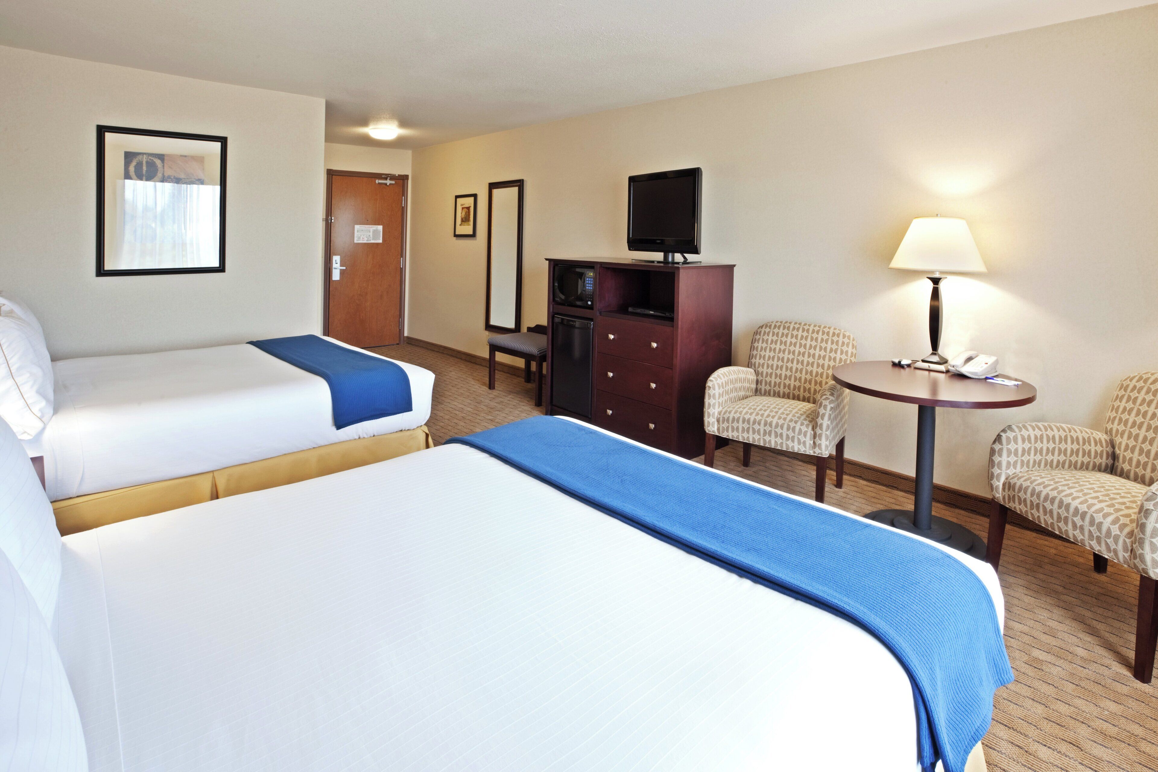 A hotel room with two beds, a television, a desk with a lamp, and two chairs.
