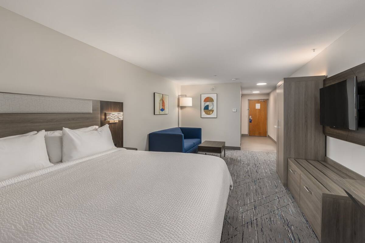 A hotel room with a large bed, a blue armchair, a flat-screen TV, and a wooden cabinet.