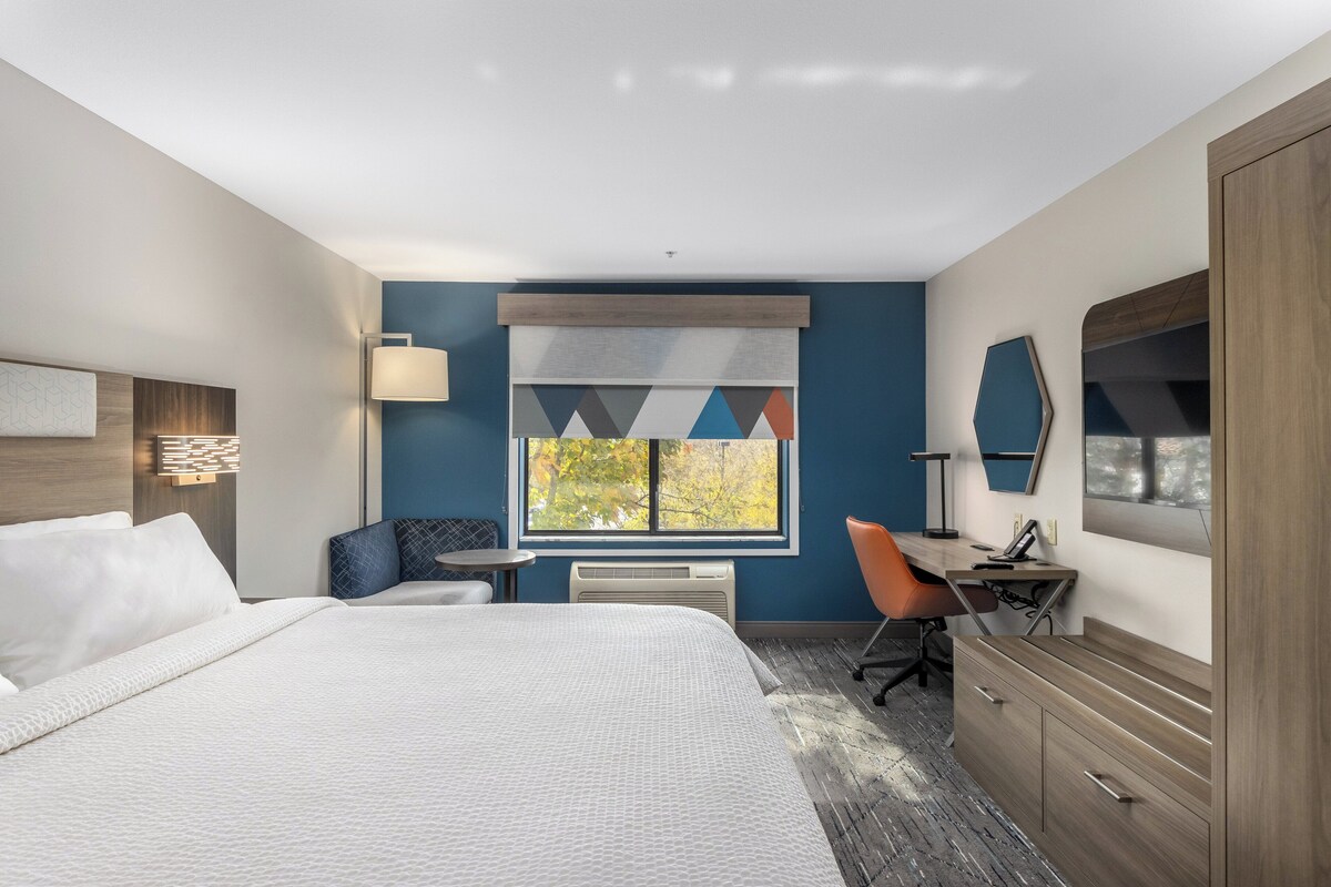 A hotel room with a large bed, a desk, a chair, a wardrobe, and a window with a view of trees.