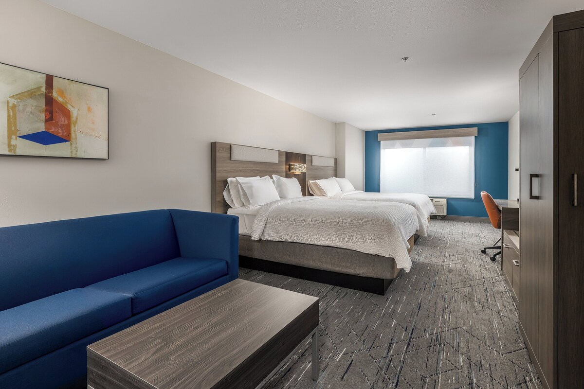A hotel room with a blue sofa, two beds, a desk, and a large window.