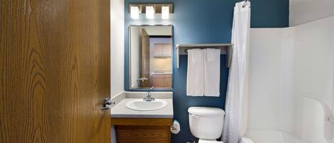 A bathroom with a wooden door, a sink, a toilet, a bathtub, and a mirror with lights above it.