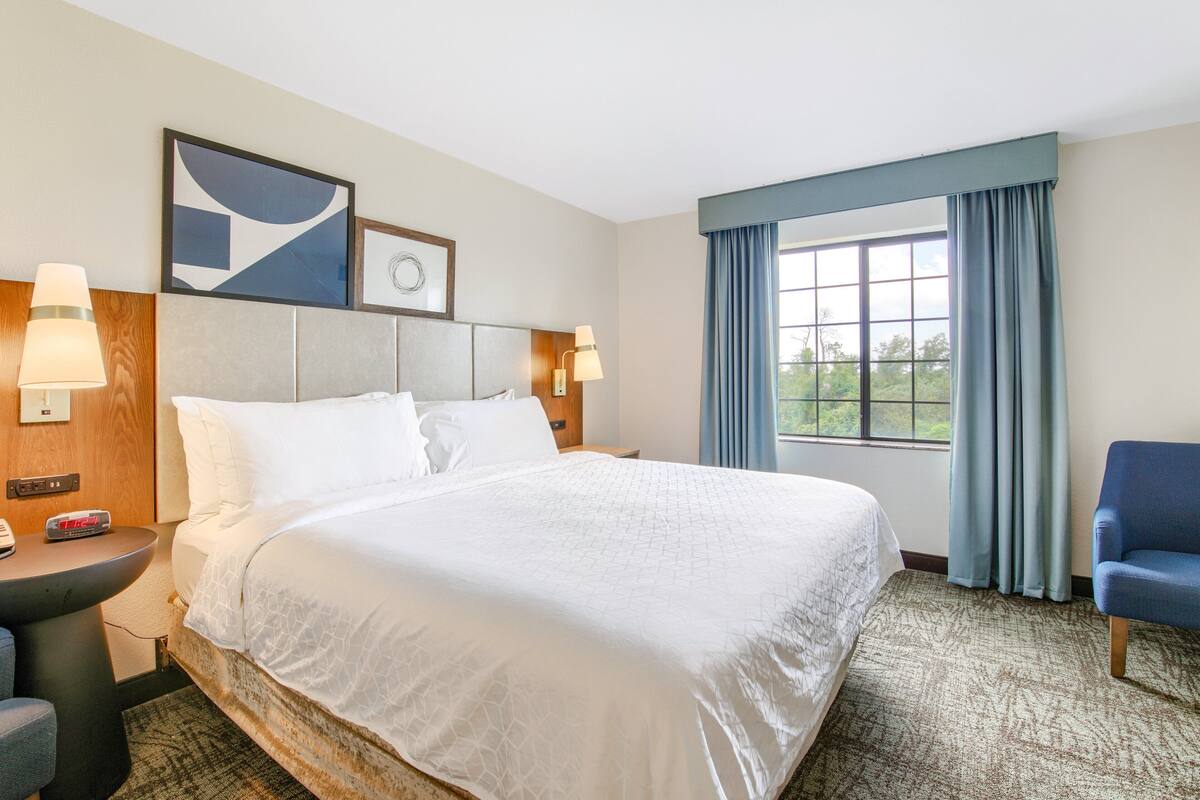 Hotelkamer met een groot bed, twee bedlampjes, een bureau met lamp, een stoel, een raam met gordijnen en een wandklok.