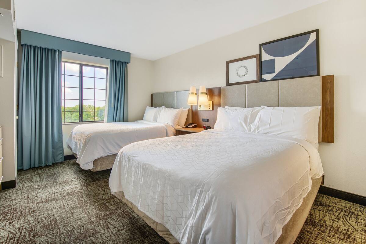 Hotelkamer met twee bedden, een groot raam met gordijnen, een hoofdeinde met wandlampen en een abstract kunstwerk erboven.