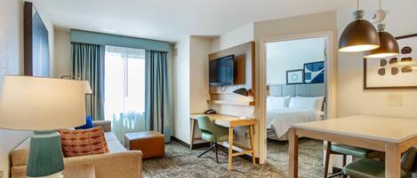 A hotel room with a sofa, a desk, a dining table, a TV mounted on the wall, and a bed in the background.