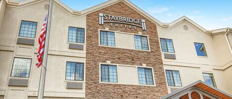 Edificio del hotel Staybridge Suites con fachada de piedra, múltiples ventanas y una bandera estadounidense.
