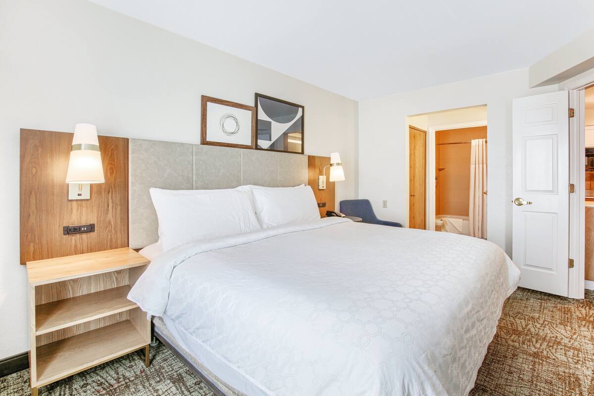 Hotelkamer met een groot bed, nachtkastjes, een bureau en een stoel. Er hangen foto’s in lijsten aan de muur en een deur die naar een andere kamer leidt.