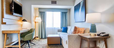Habitación de hotel con televisor de pantalla plana, escritorio con silla, sofá con cojines, mesita de noche con lámpara y ventana grande con cortinas.