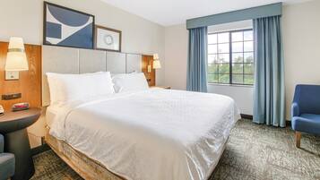 Habitación de hotel con una cama grande, dos lámparas de noche, un escritorio con lámpara, una silla, una ventana con cortinas y un reloj en la pared.