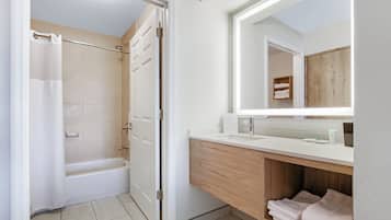 A bathroom with a bathtub, a shower curtain, a vanity with a mirror, and a shelf with towels.