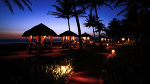 A beachside resort at dusk with thatched-roof cabanas, palm trees, and a pathway leading to the beach.