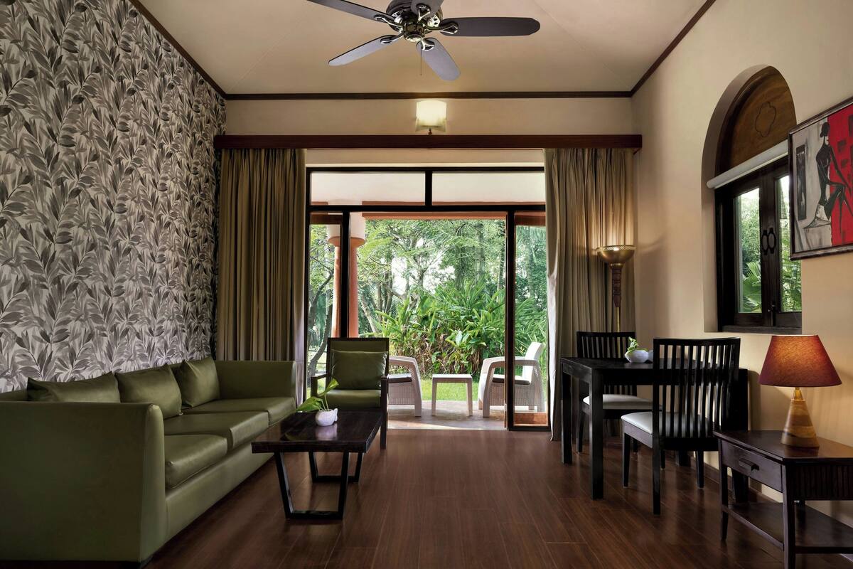 Una habitación con sofá verde, una mesa de centro, una mesa de comedor con sillas, un ventilador de techo y vistas a la vegetación a través de la puerta de cristal.