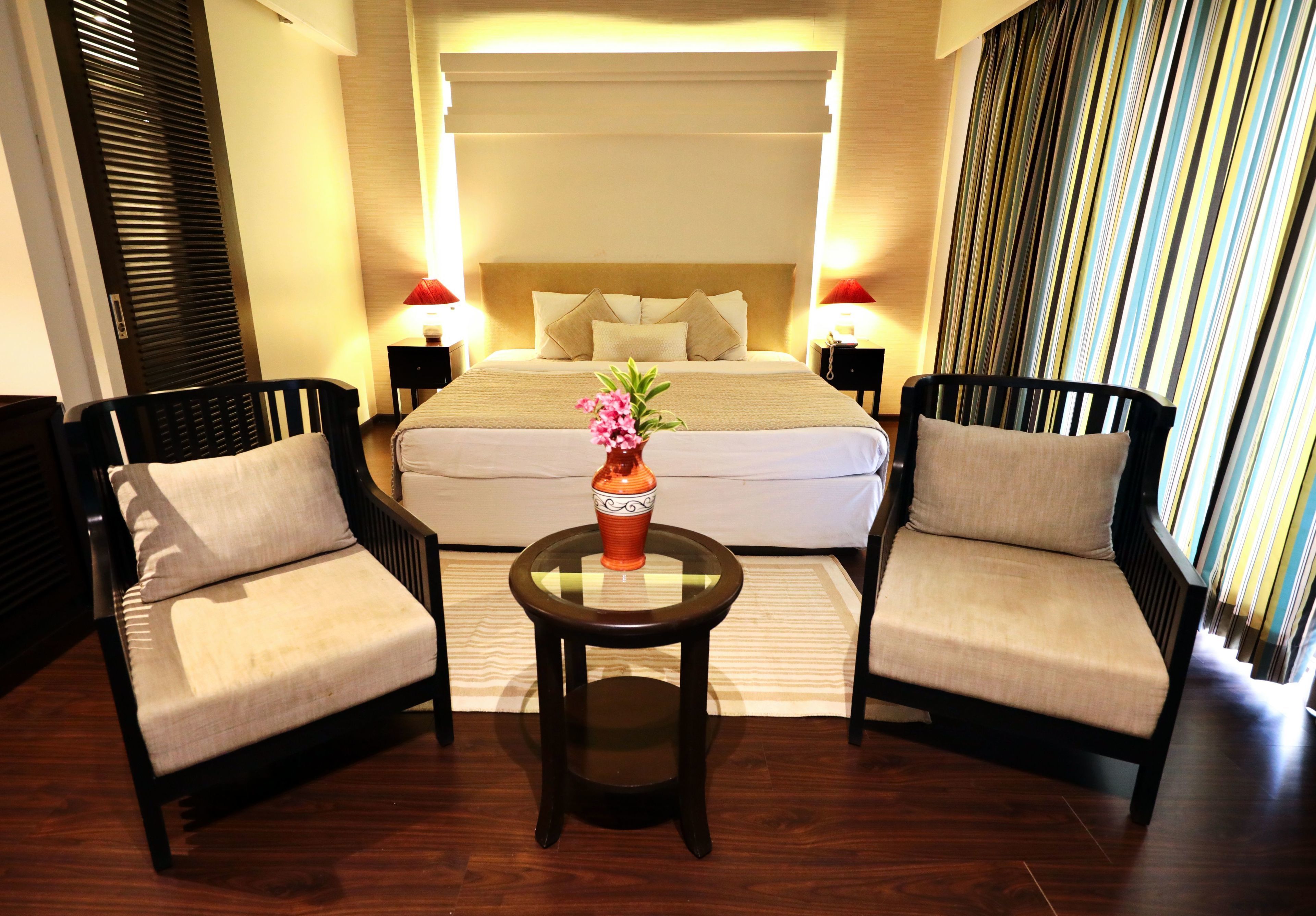 Una habitación de hotel con una cama grande, dos sillones, una mesita con un jarrón y dos lámparas de noche.