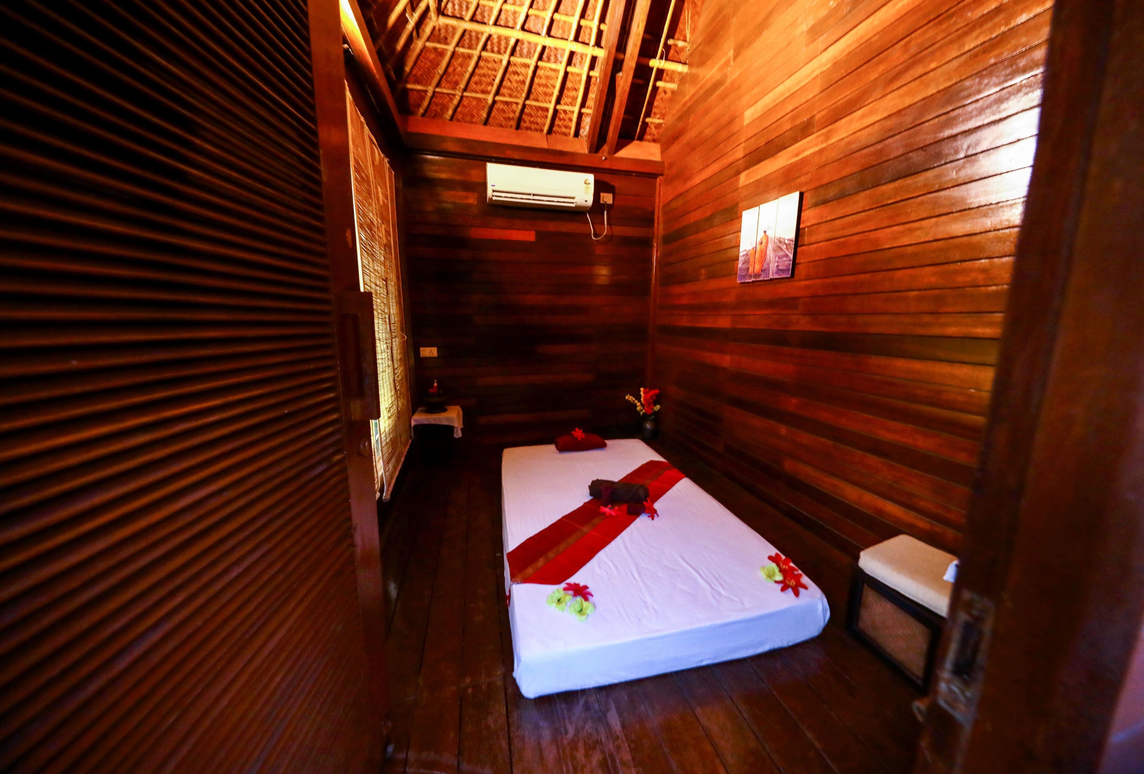 Quarto pequeno e compacto com paredes de madeira e teto inclinado. Possui uma cama de solteiro com colchão branco, uma mesinha de cabeceira e um ar condicionado fixado na parede.