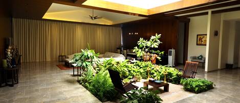 A spacious living area with a high ceiling, a skylight, and a variety of plants arranged around a central seating area.