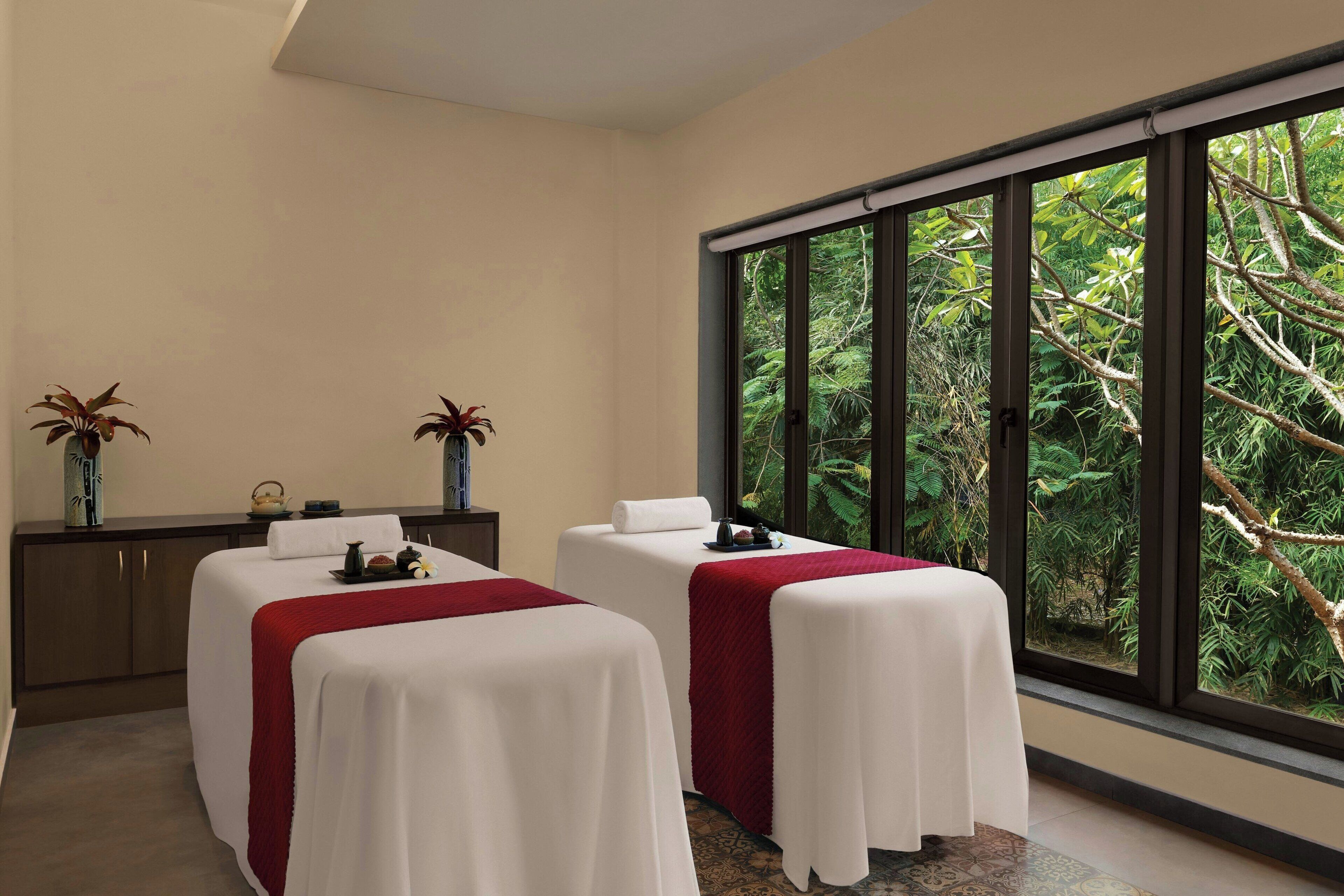 A room with two massage tables covered in white sheets and red blankets, a wooden cabinet with decorative items, and a large window offering a view of lush greenery.