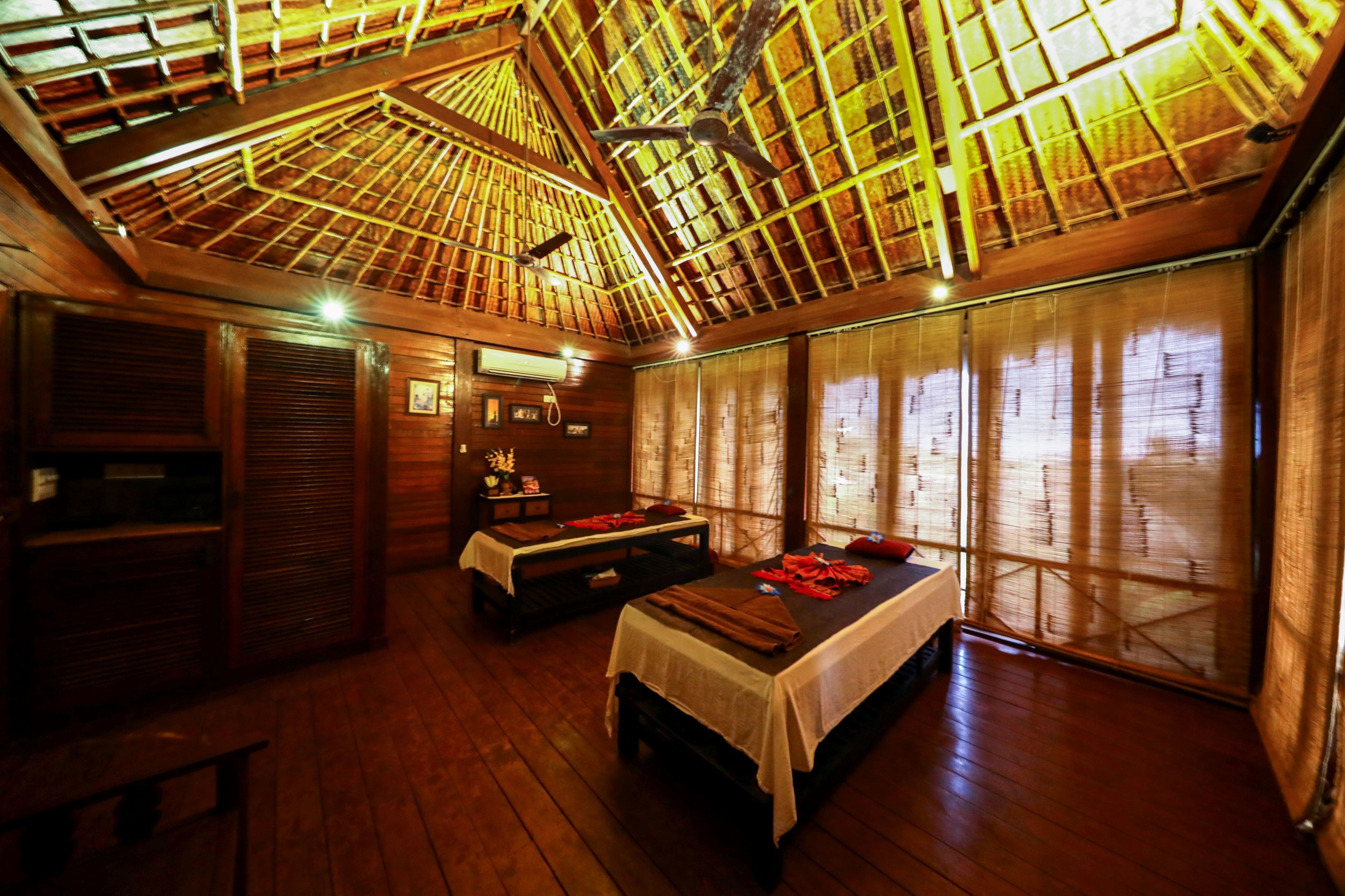A room with a wooden ceiling, two beds, a bench, and a window with bamboo blinds.
