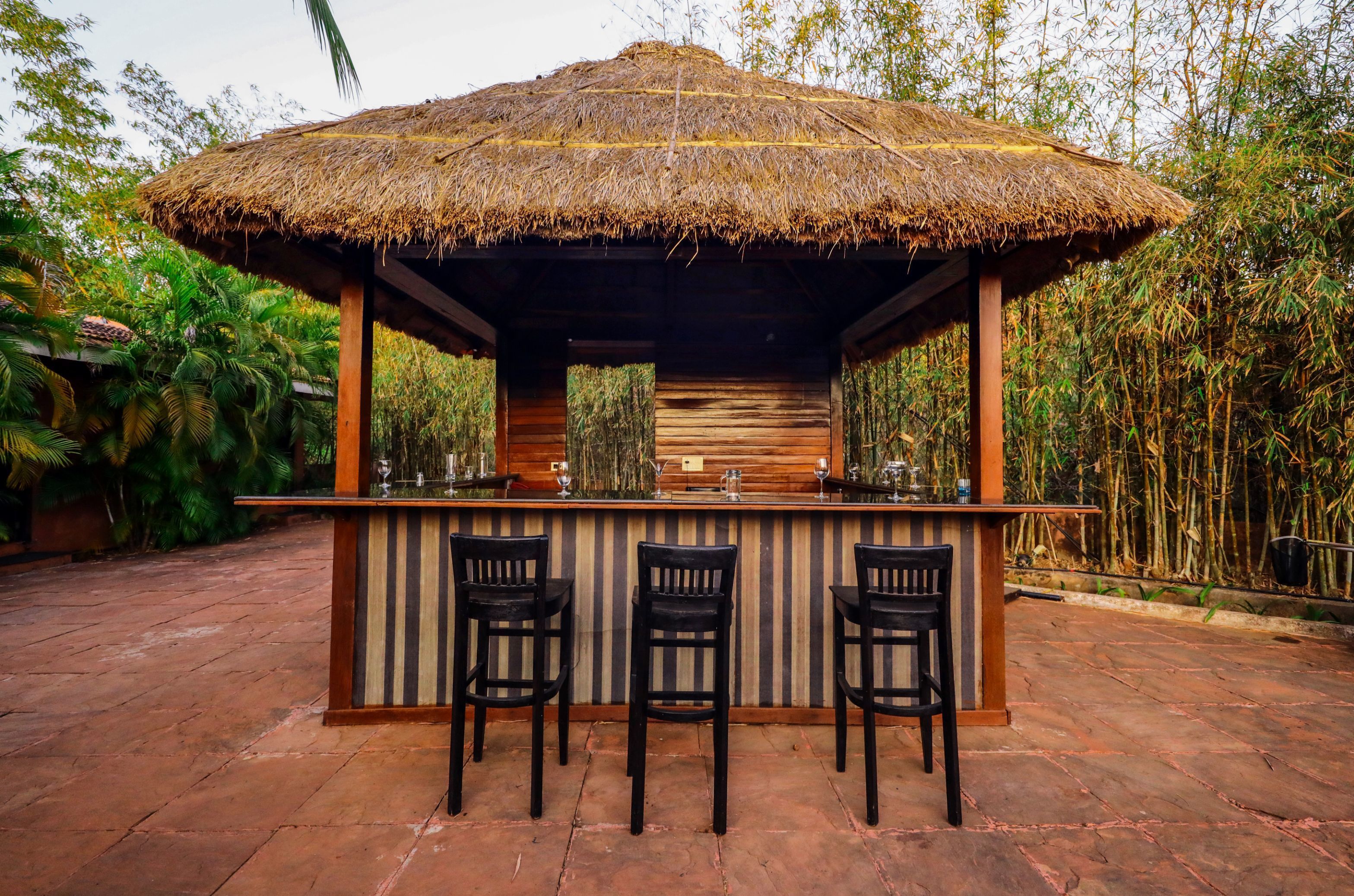 A thatched-roof outdoor bar with three wooden chairs, surrounded by lush greenery.