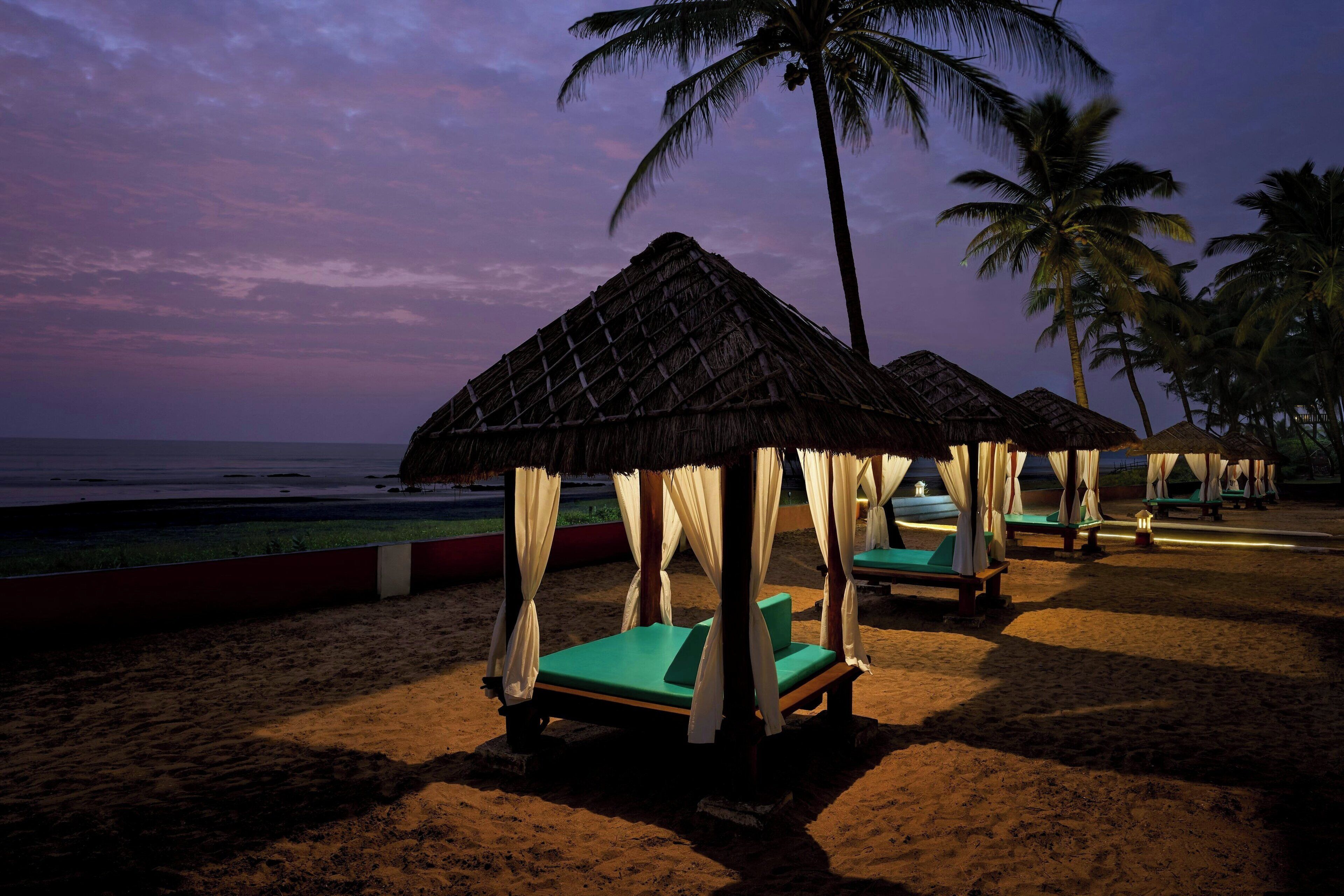 Een strand met rieten hutten, groene lounge stoelen en palmbomen.