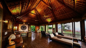 A spacious room with a wooden ceiling, a reception desk, a sofa, and large windows offering a view of the outdoors.