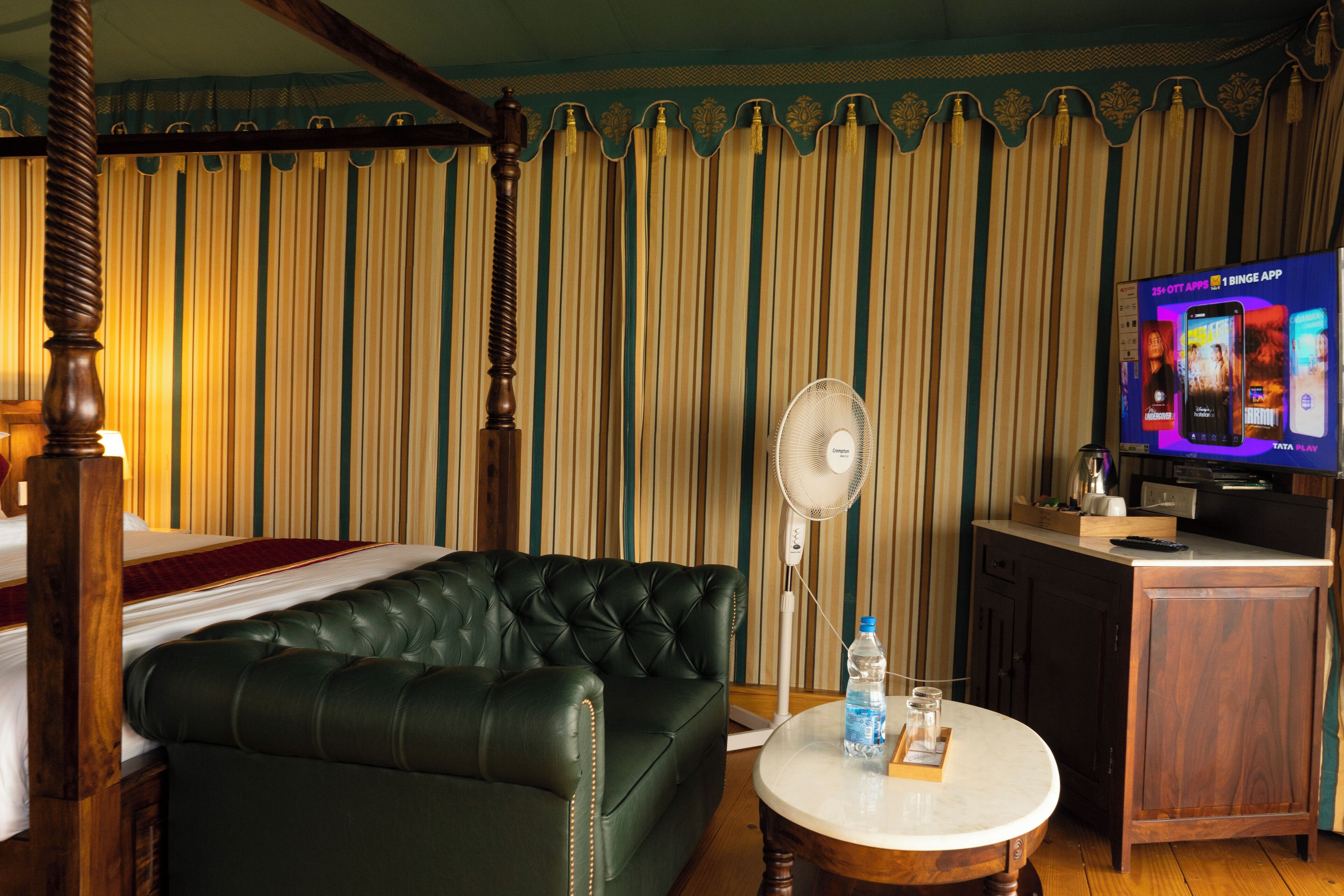 A hotel room with a four-poster bed, a green leather sofa, a round wooden table with a bottle and glass, a standing fan, and a TV displaying an advertisement.