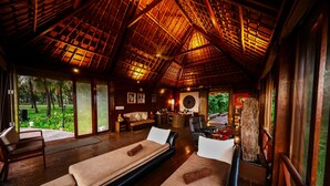 A room with wooden flooring, a thatched roof, and a large window overlooking a garden. There are two massage tables with towels and a sofa.