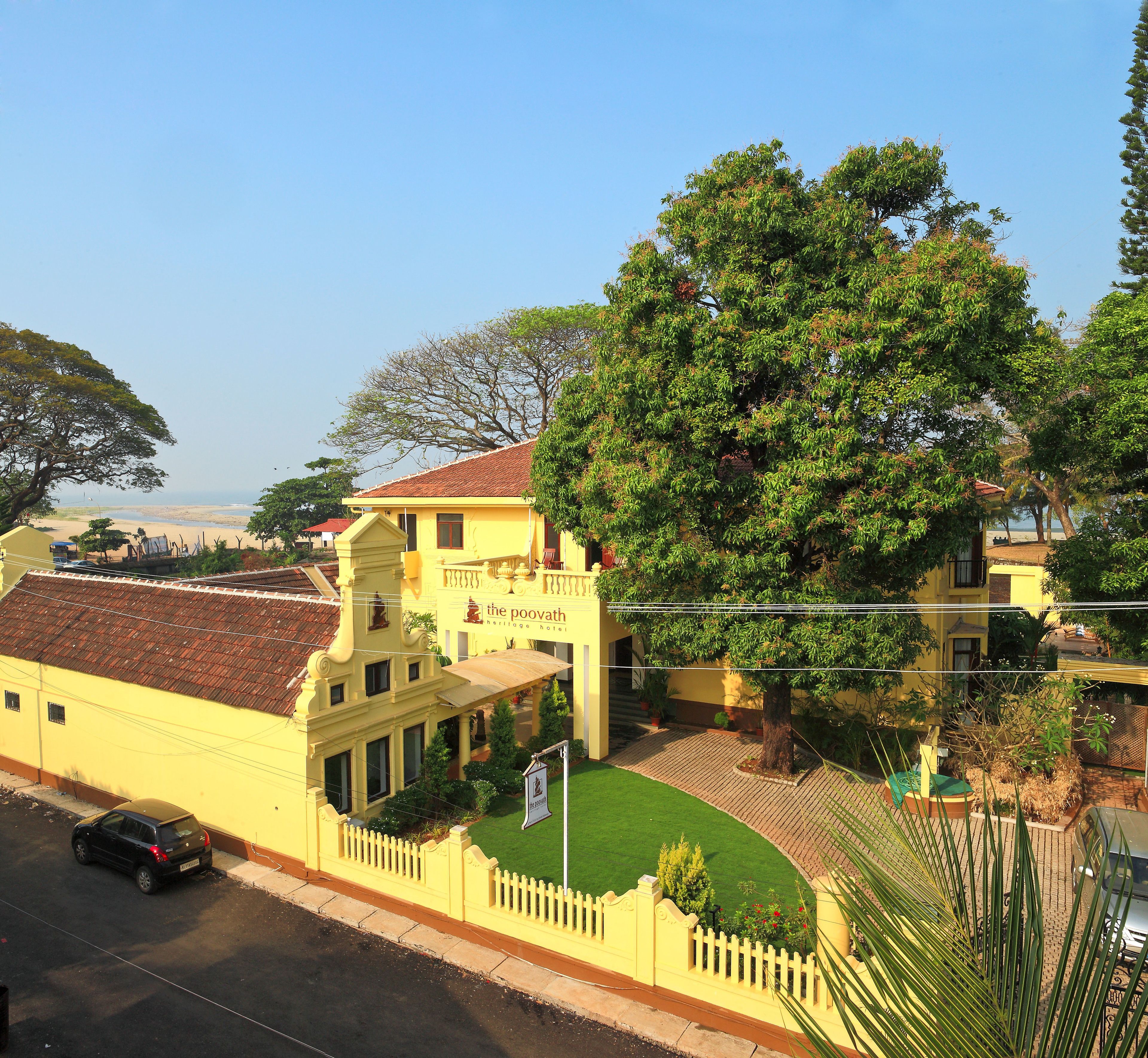 A yellow heritage hotel with a red-tiled roof, a large tree in front, and a view of the beach.