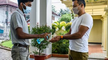 Deux hommes échangent un petit objet, debout sous une zone abritée, entourés de plantes en pot, avec un bâtiment en arrière-plan.
