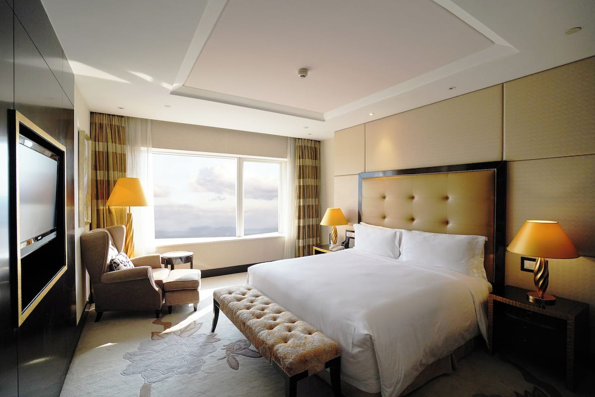 A hotel room with a large bed, a flat-screen TV, a seating area with a chair and bench, and a window with curtains.