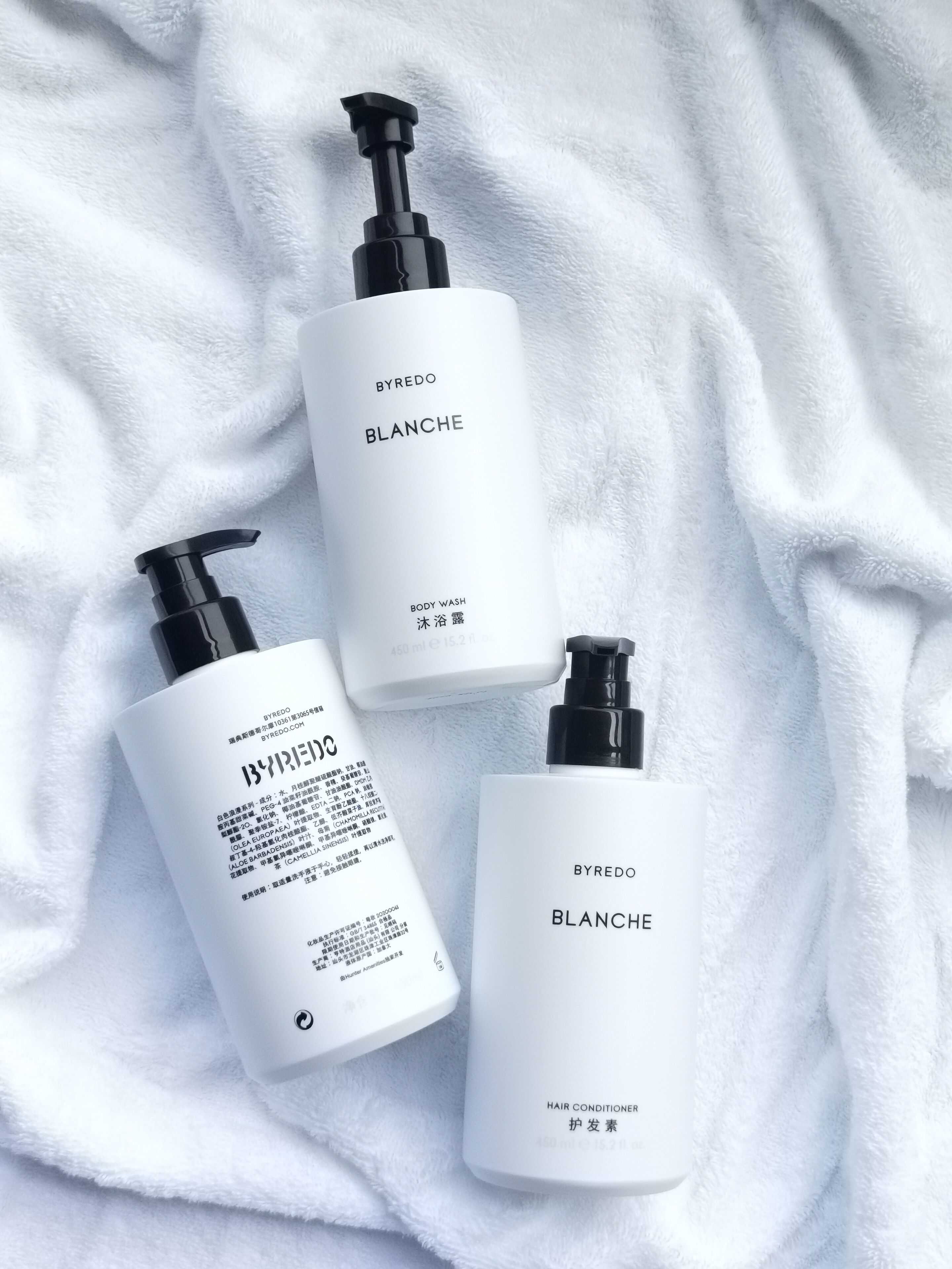 Tres botellas blancas de gel de ducha y acondicionador para el cabello BYREDO BLANCHE sobre una toalla blanca.