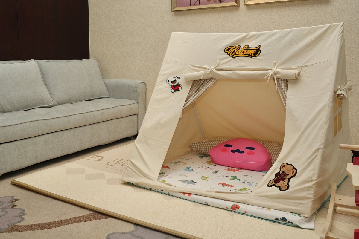 Ein gemütliches Zimmer mit einem grauen Sofa, einer kleinen, zeltartigen Spielgelegenheit mit einem rosa Kissen und einem gemusterten Teppich.