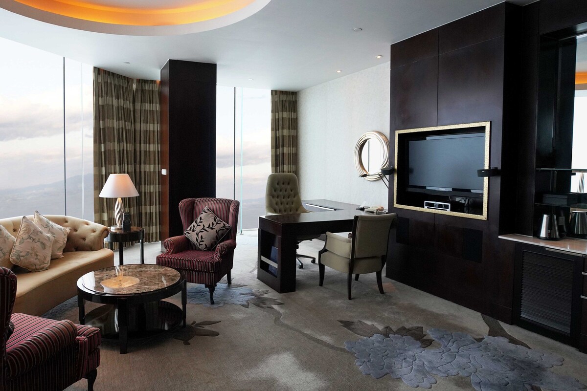 Ein modernes Hotelzimmer mit großem Fenster, einer Couch, einem Sessel, einem Schreibtisch und einem Fernseher.