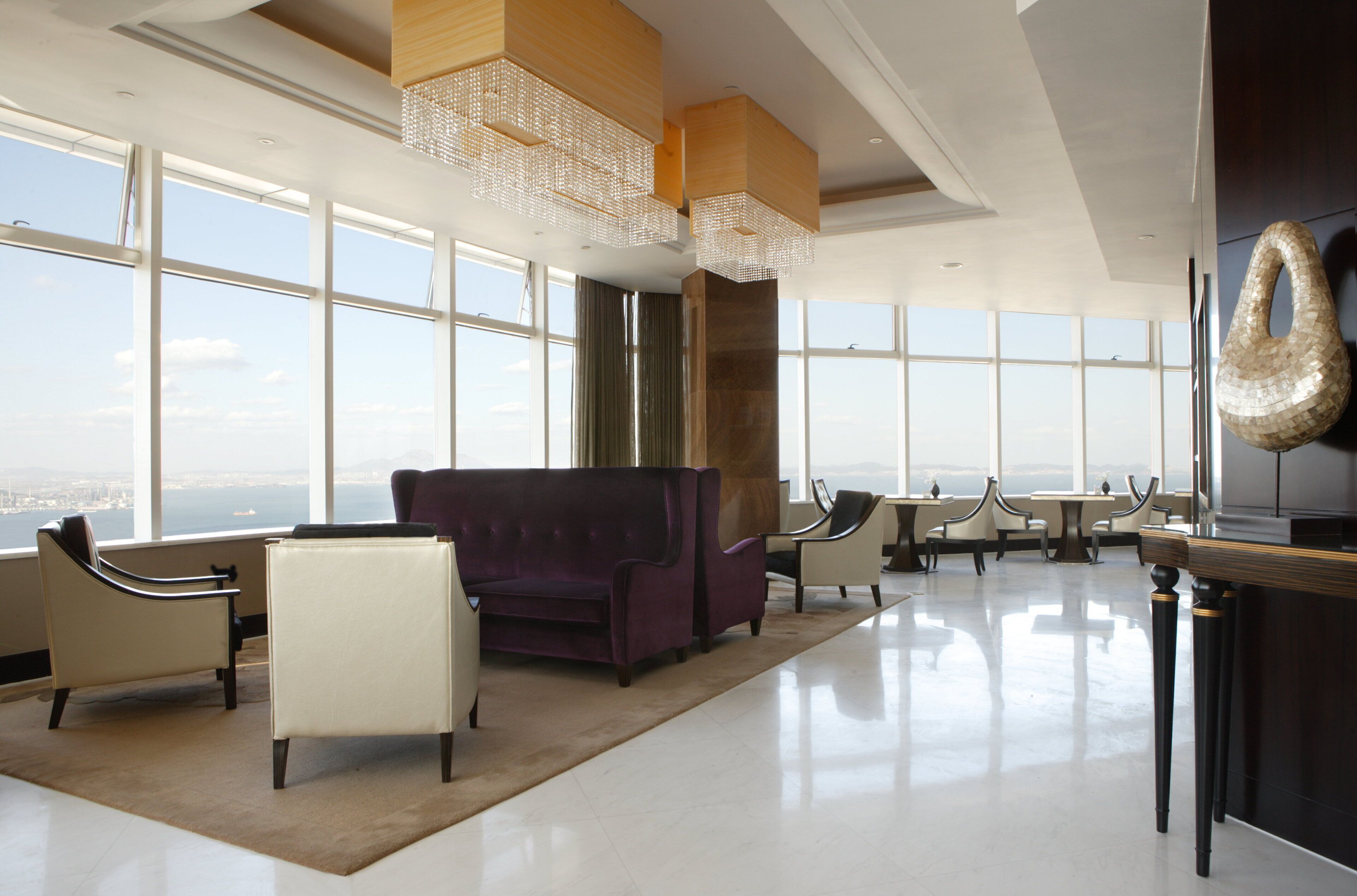 Un'ampia zona relax con una grande finestra che si affaccia sulla città, un divano viola elegante e un lampadario moderno.