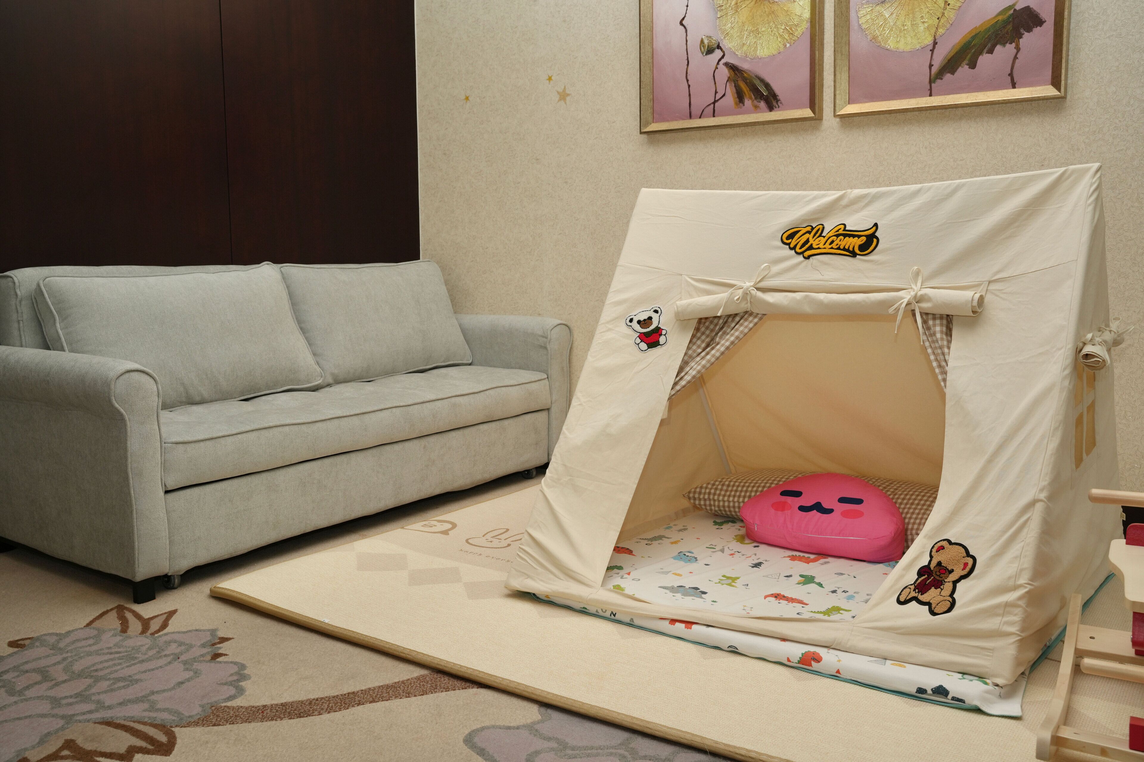 Une pièce avec un canapé gris, un petit espace de jeu aménagé comme une tente avec un coussin rose, et un tapis à motifs floraux.